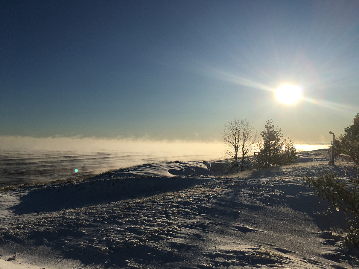 Frost smoke rising above the surface of Lake Superior with snow-covered dunes in the foreground and a bright sun above the horizon