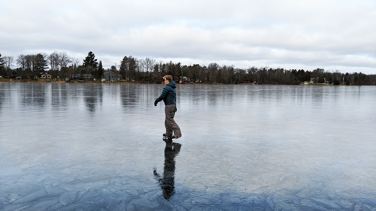 Youth skating on lake covered in clear, patterned ice. Tree-lined shore in the background. Slightly overcast, cloudy sky. 