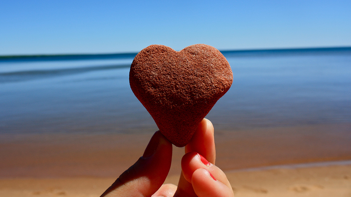 Heart-shaped rock held by two fingers with Lake Superior beach and water in background.