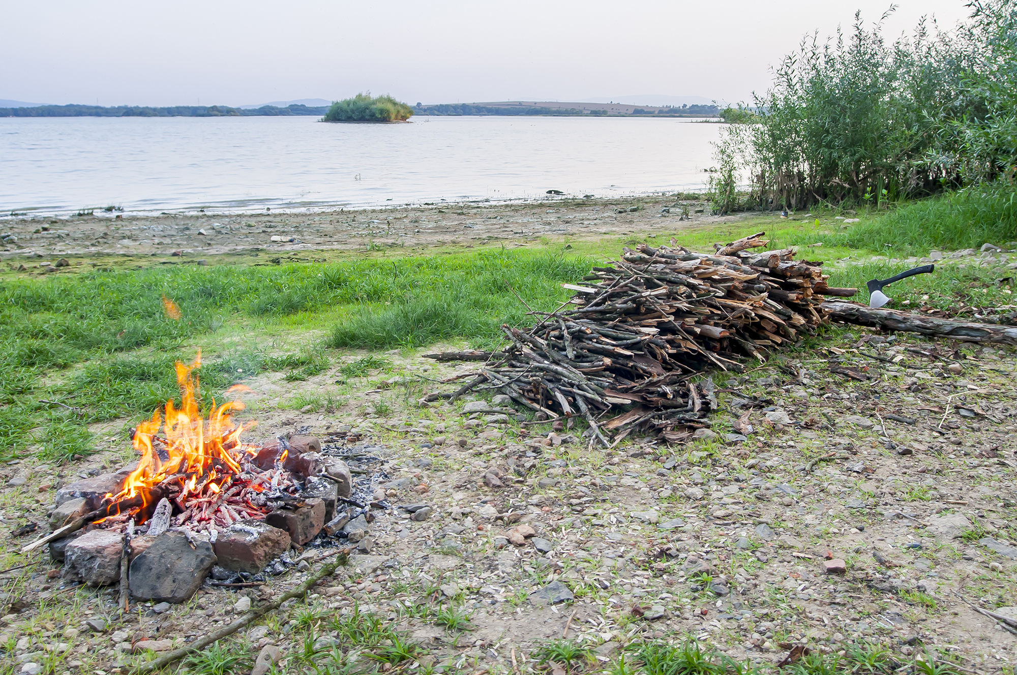 Small campfire with flames beside a lake with wood pile on the side.