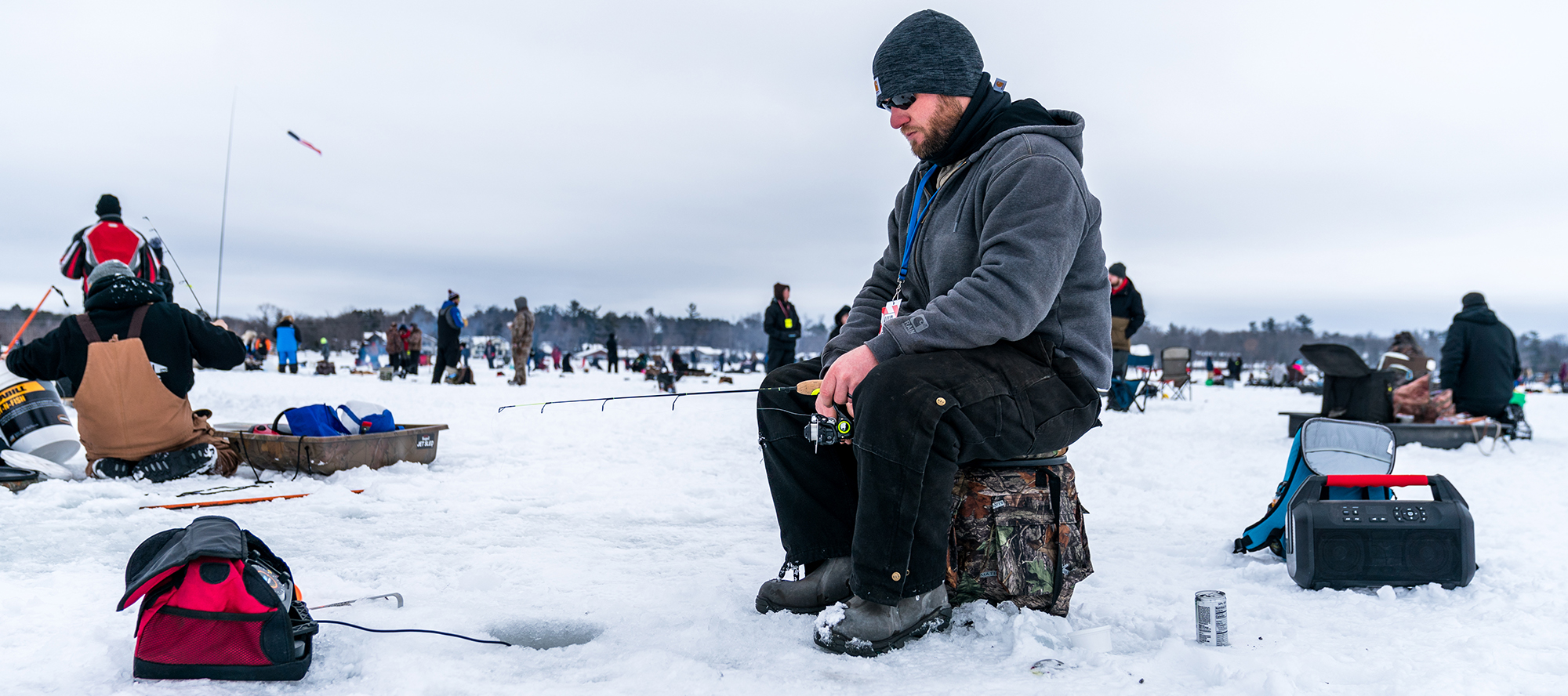 Man sitting watching ice-fishing hole on a snow-covered lake, many people in background.