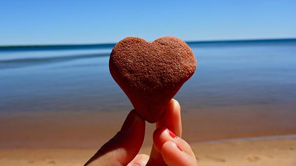 Heart-shaped rock held by two fingers with Lake Superior beach and water in background.