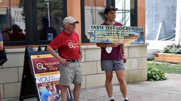 Two people standing on sidewalk, one holding a sign that reads: taste test local fish.