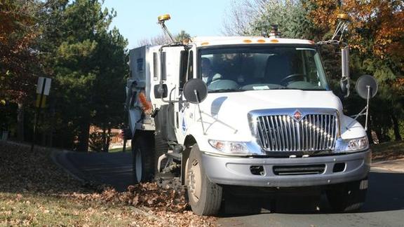A white street sweeper picks up fallen leaves off a roadway.