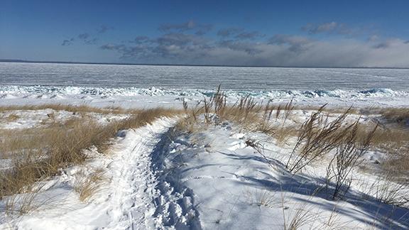 Snow-covered sand dune with foot-trodden path leading to ice-covered lake.