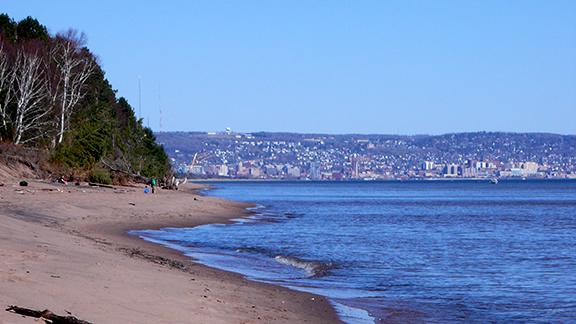 Picture of a brown, sandy beach and tree line to the left with blue lake water to the right and a cityscape in the background.