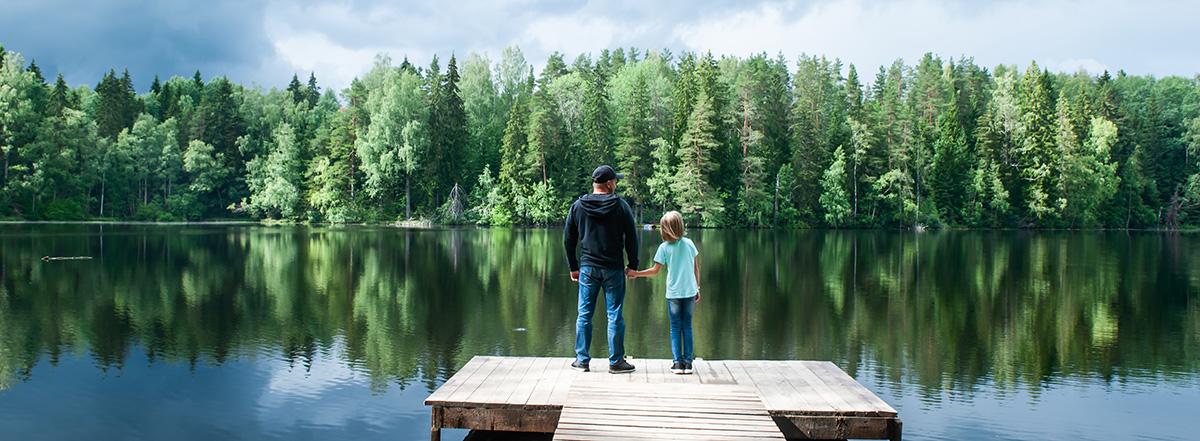 Two people holding hands standing on a wooden dock looking out toward a lake and a forest.