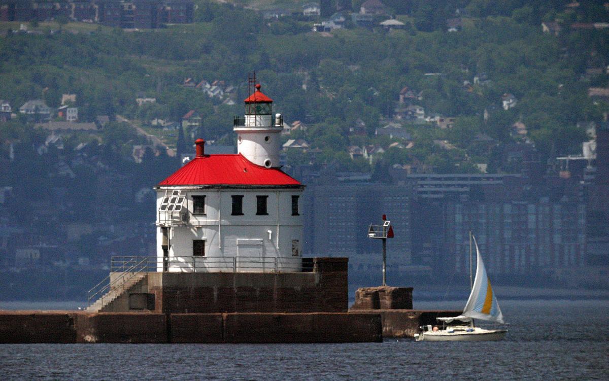 Red and white lighthouse at end of pier, sailboat in water below, city skyline in distance.