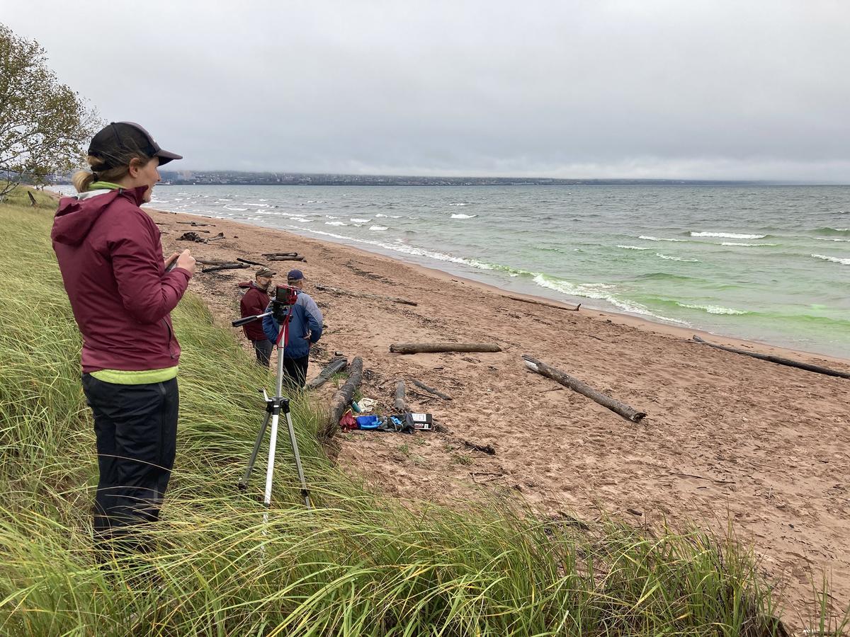 Person standing on dune near camera, two people standing on sandy beach. All three people looking toward Lake Superior and watching the movement of food-grade green dye in the water used to visualize a rip current.