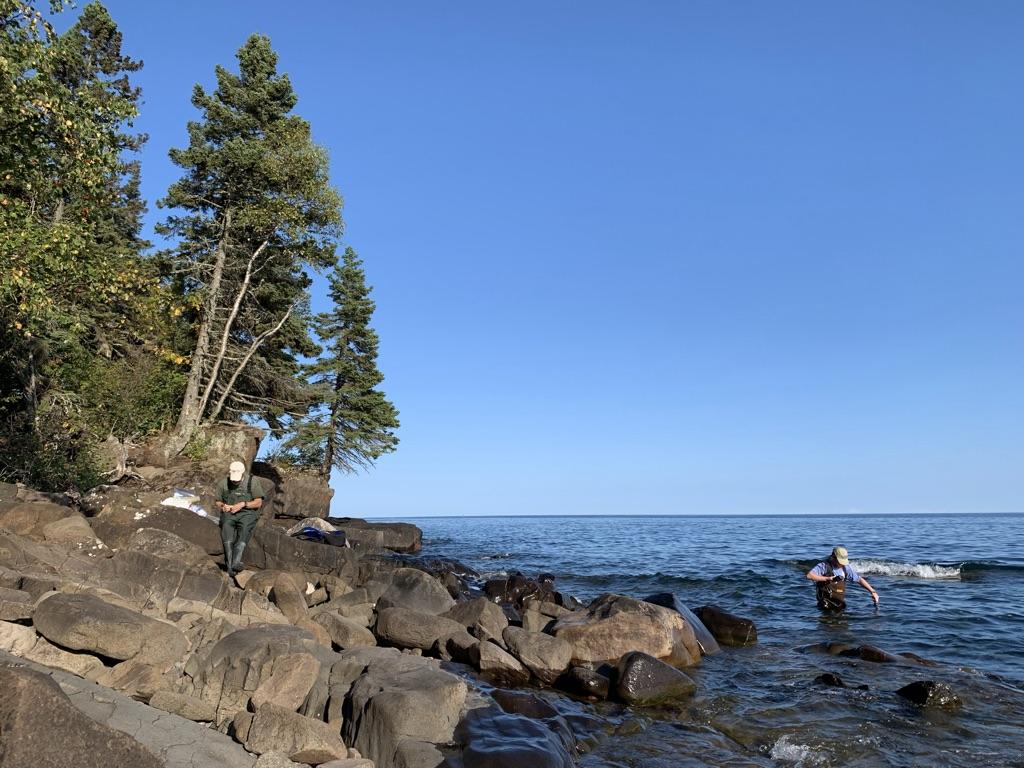 Rocky shore of Lake Superior near the Cascade River. One person in waders in the nearshore water. One person sitting on the rocky shore. 