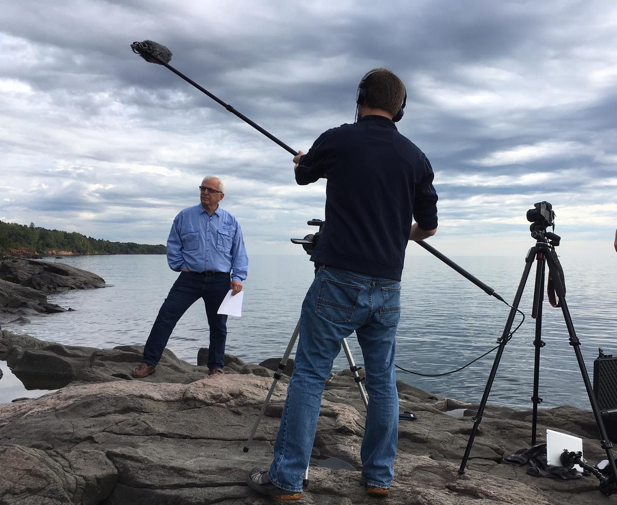 Two people on rocky shoreline. One person with a recording boom microphone.
