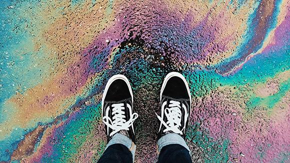 Top view of a person's feet in shoes, standing in an oil spill on pavement.
