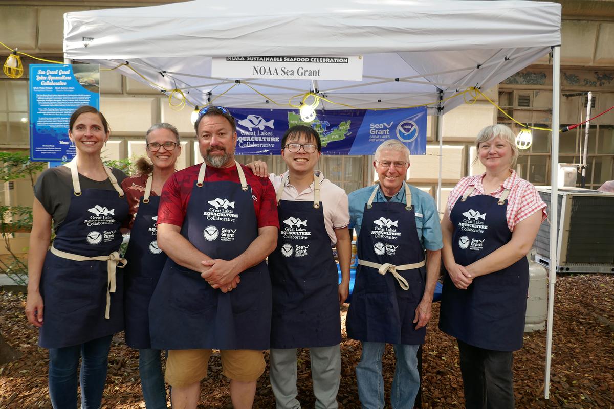 Six people standing together in front of a booth wearing aprons. 