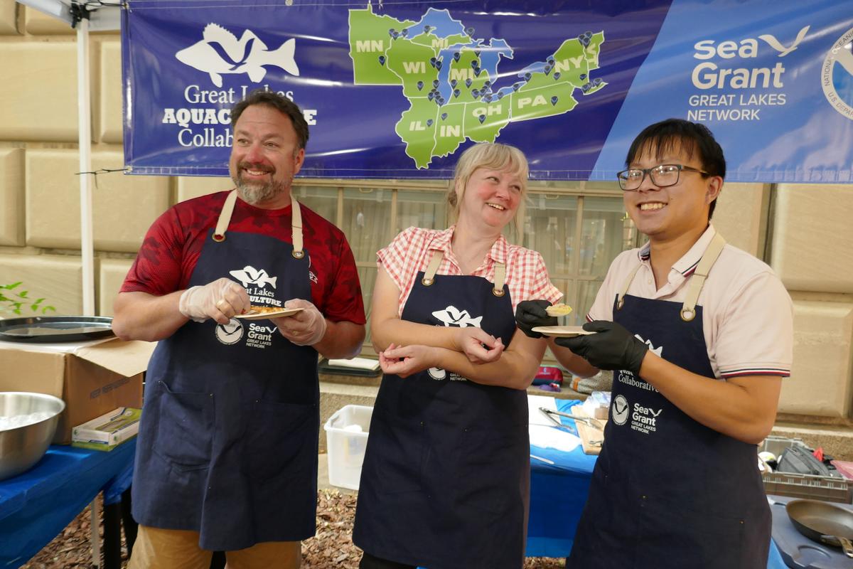 Three people wearing aprons and sampling seafood.
