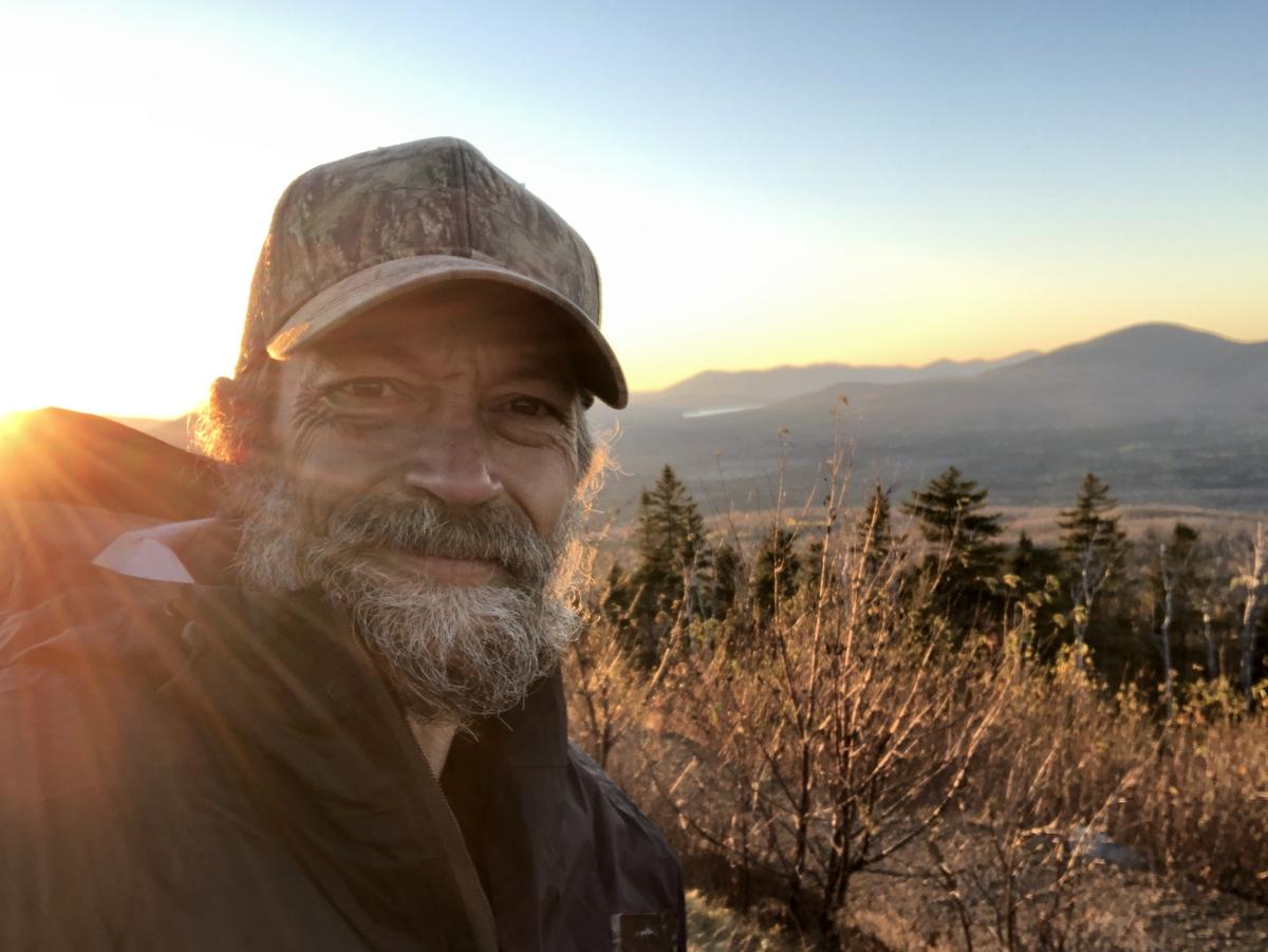 Erik Simula smiles in front of a sunset over forested Appalachian mountains in Maine.