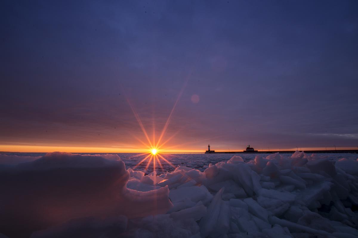 A burst of sunshine rising over an icy Lake Superior, with the silhouette of the Duluth Entry lighthouses visible.