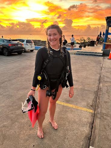 Malin Anderson smiles against a sunset while standing in a parking lot wearing a wetsuit and holding flippers.